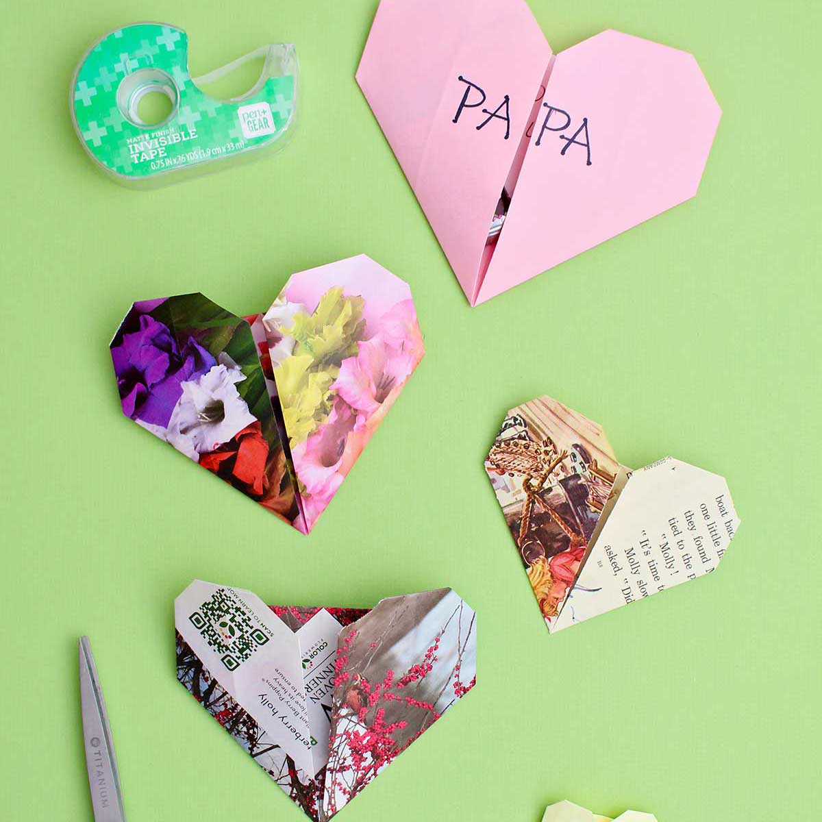Four origami hearts on a green background with scissors and tape near by.