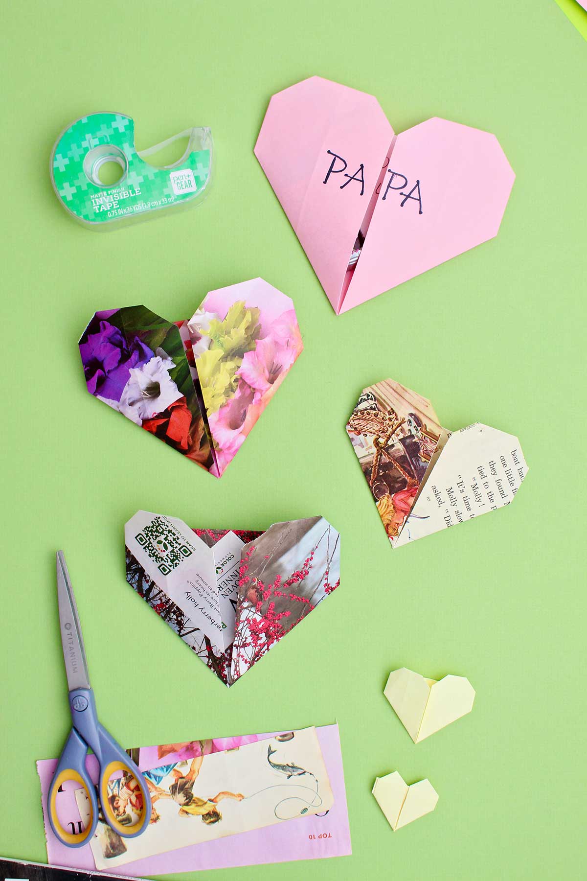Four origami hearts on a green background with scissors and tape near by.