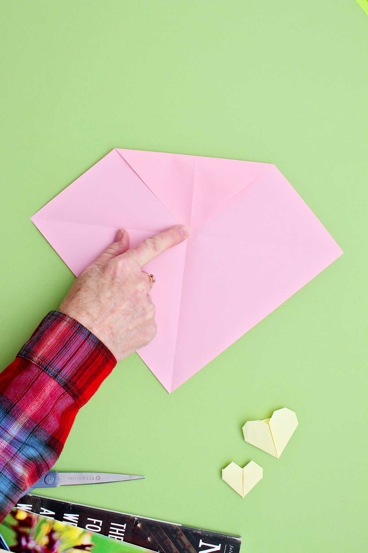 Folding a top corner of a pink piece of paper down to center to create the top part of a heart.