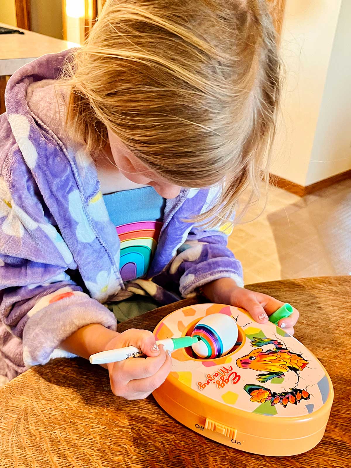 A young girl with blonde hair and a purple flowered robe coloring an egg at a kitchen table.