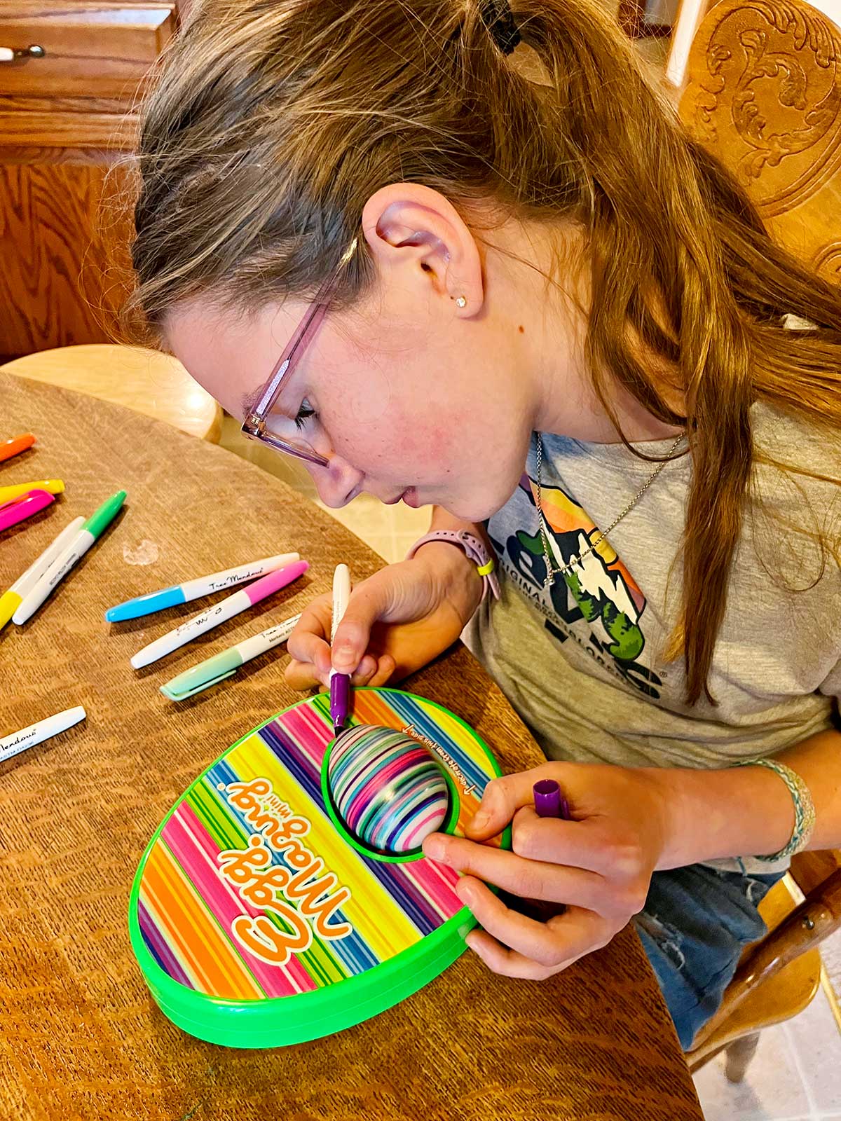 A middle school aged girl with light brown hair coloring an egg with an EggMazing machine at a kitchen table.