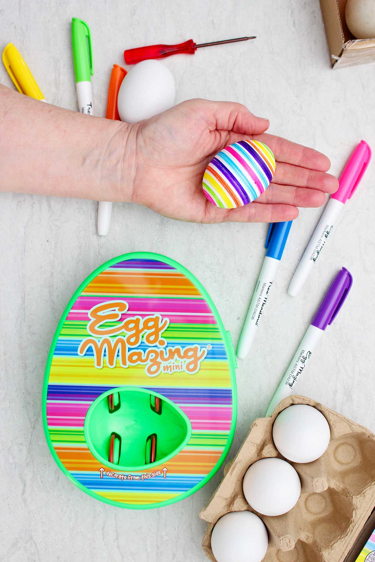 A person holding a striped colored egg next to the striped EggMazing kit.