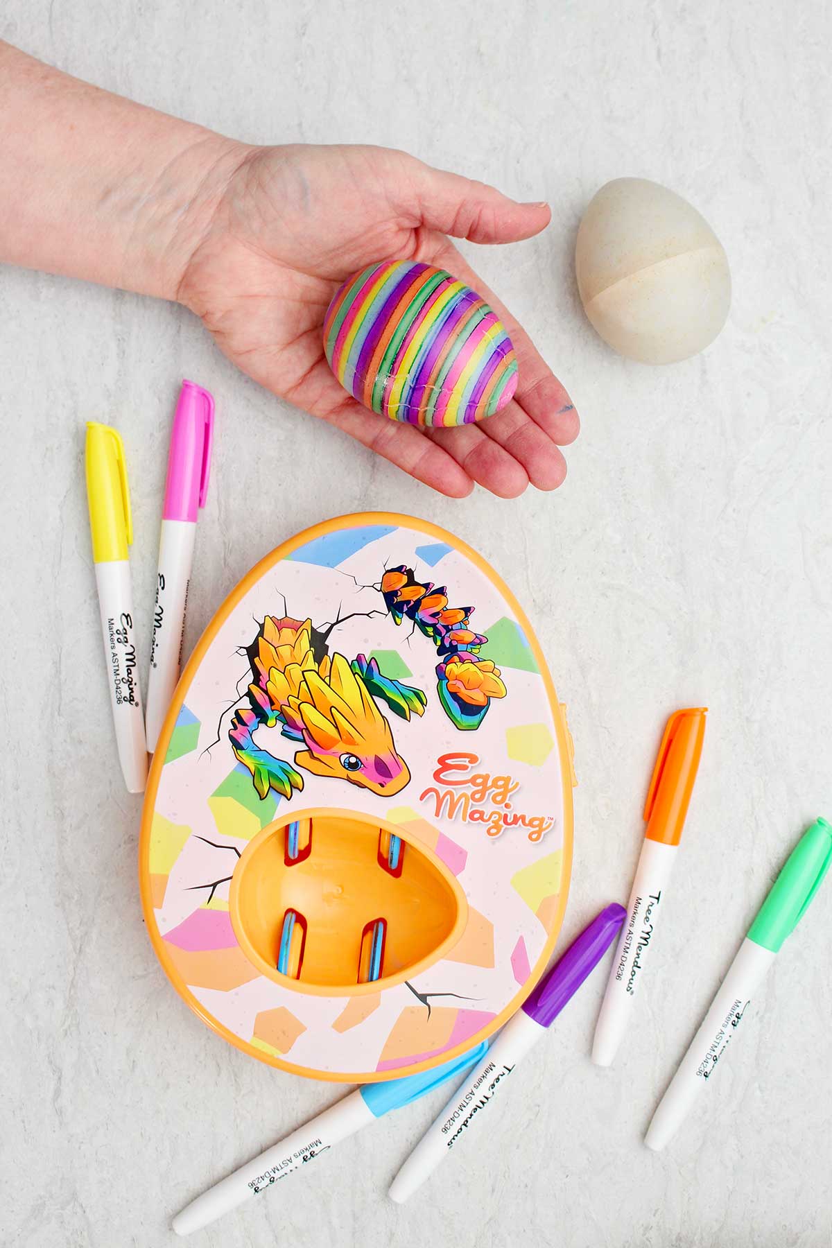 A person holding a striped colored egg with the orange dragon EggMazing kit next to them.