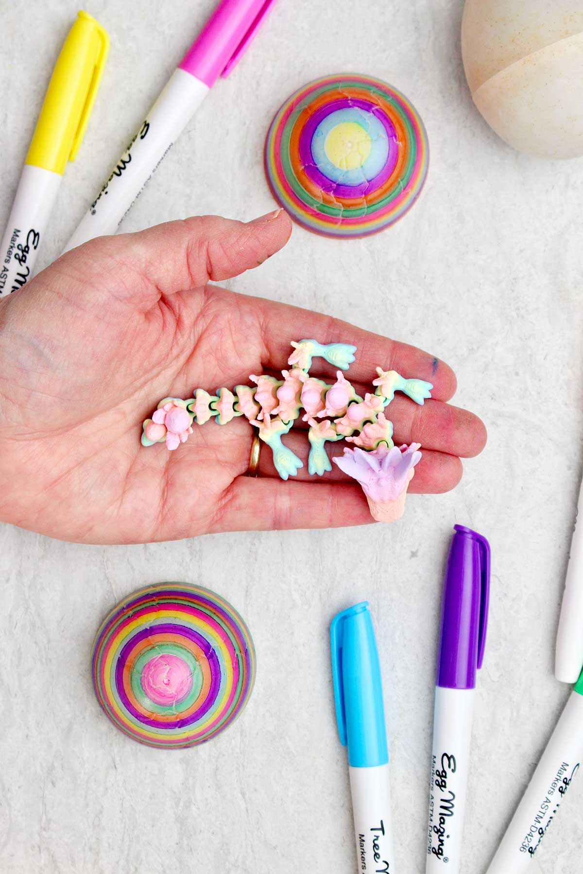 A person holding a colorful 3D printed dragon with two colored eggs and markers near by.
