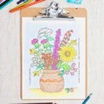 A coloring page of a brown vase full of colorful wildflowers clipped on a clipboard with colored pencils scattered around.