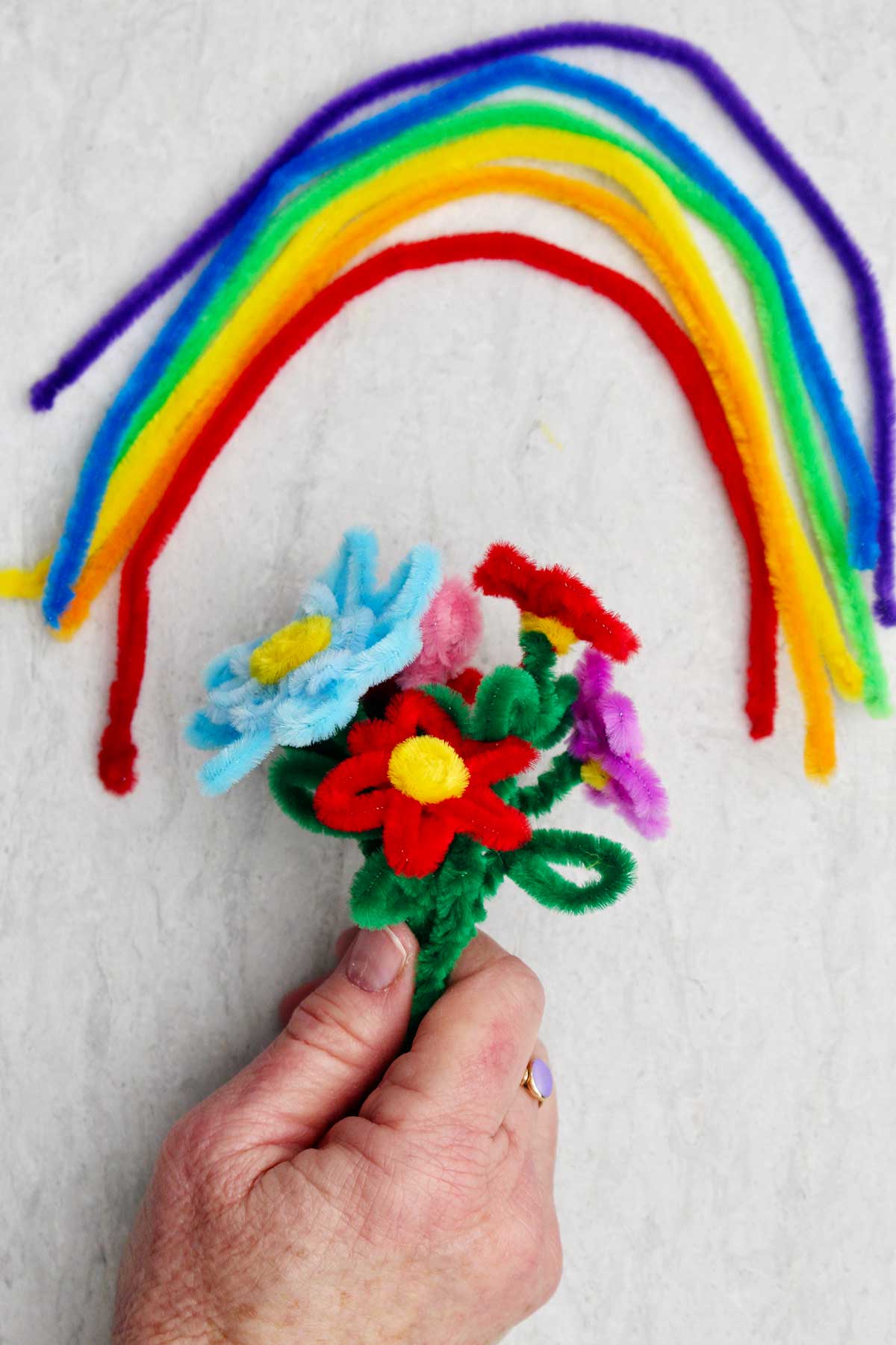 A person holding a bouquet of different colored pipe cleaner flowers with pipe cleaner rainbow above it.