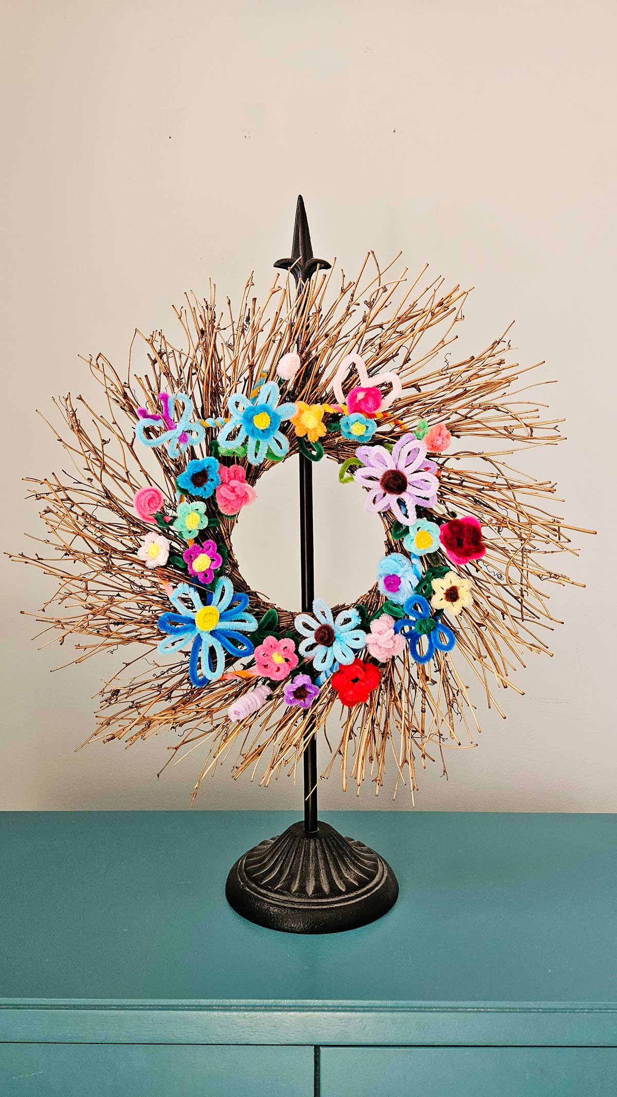 A stick wreath on a stand with multicolored pipe cleaner flowers attached to it.
