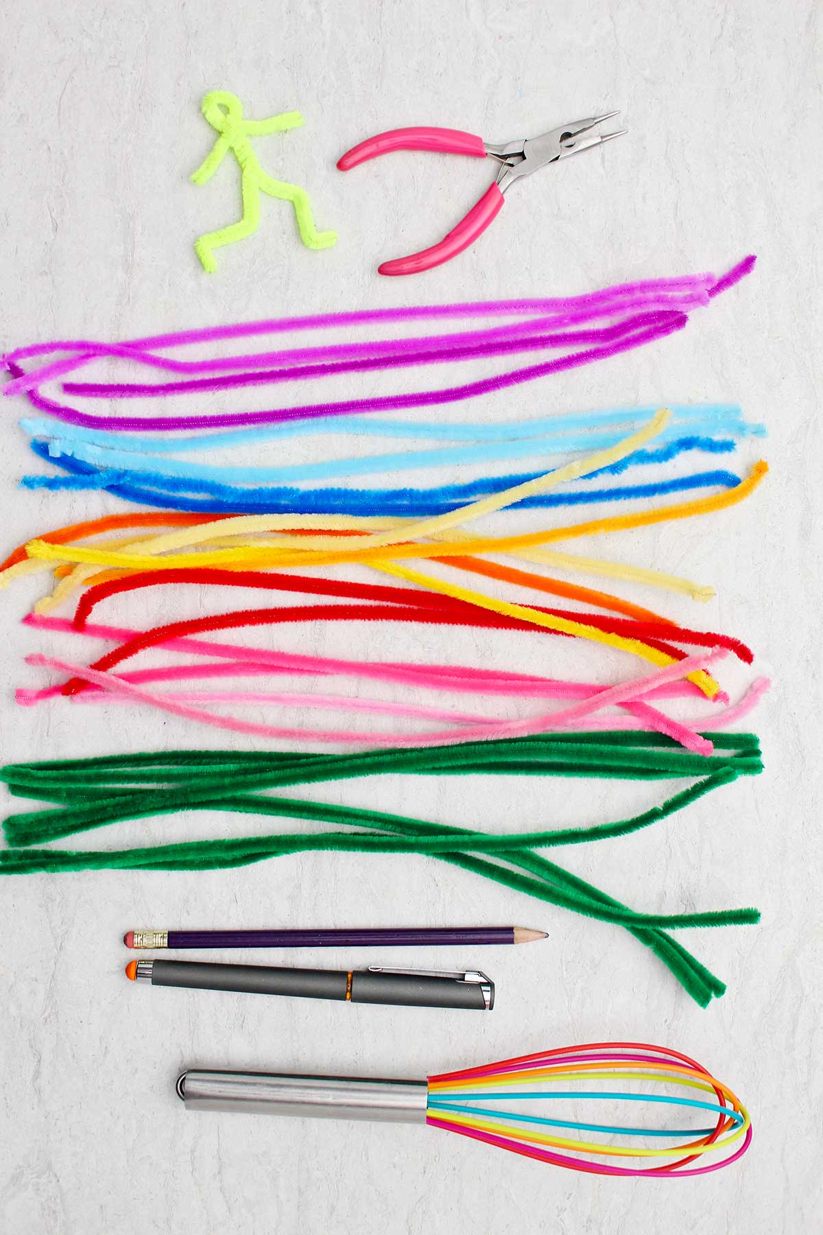 A bunch of colorful pipe cleaners, a multi colored whisk, a pen, pencil and needle nose pliers.