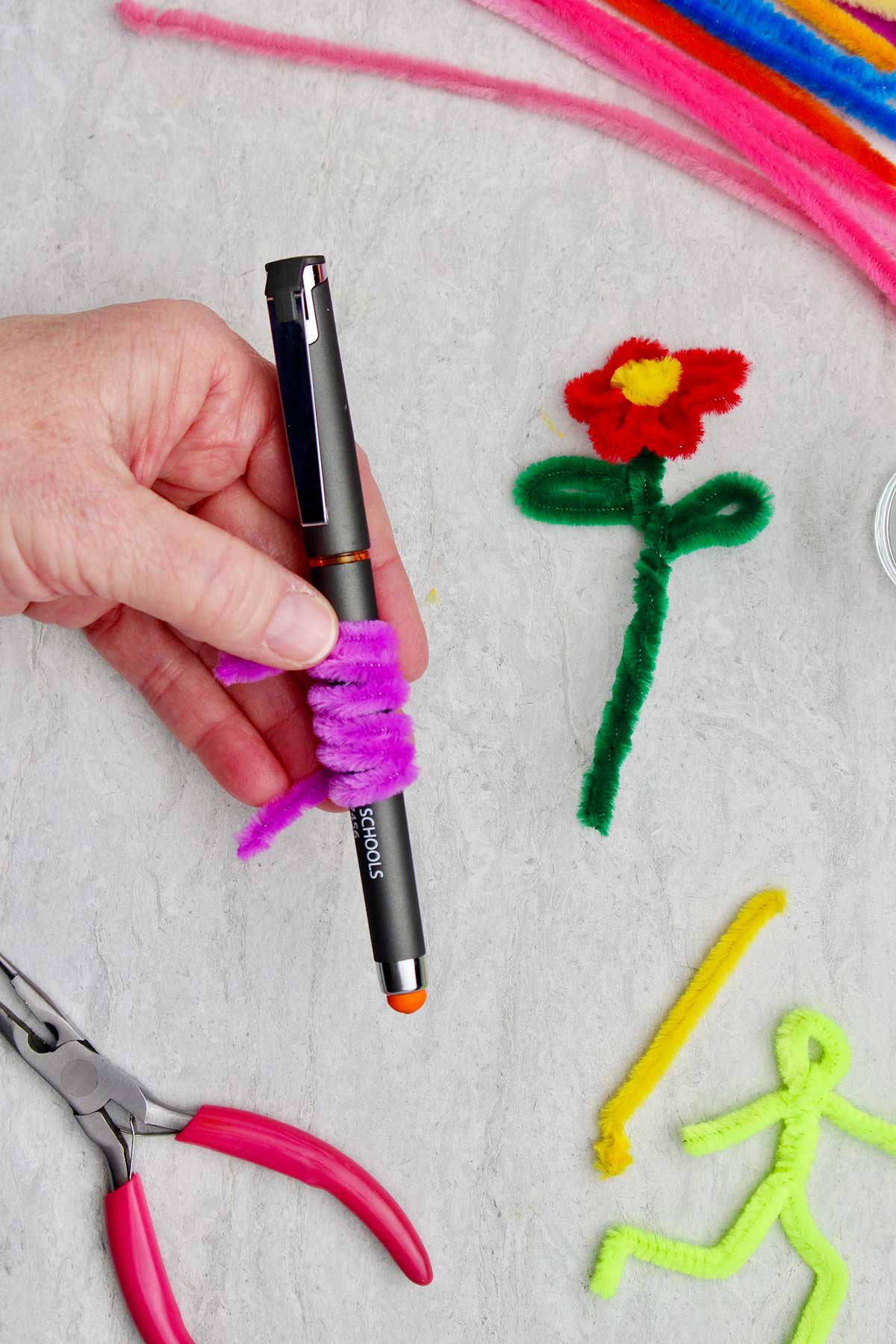 A person holding a pen with a purple pipe cleaner wrapped around it with a pipe cleaner flower and other pipe cleaners in the background.