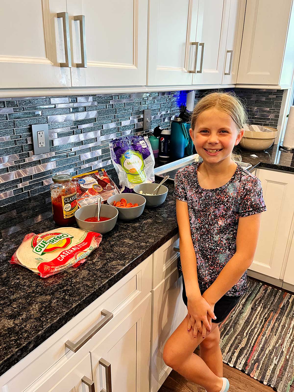 A young girl posing in front of all the supplies needed for a pizza quesadilla in the kitchen.