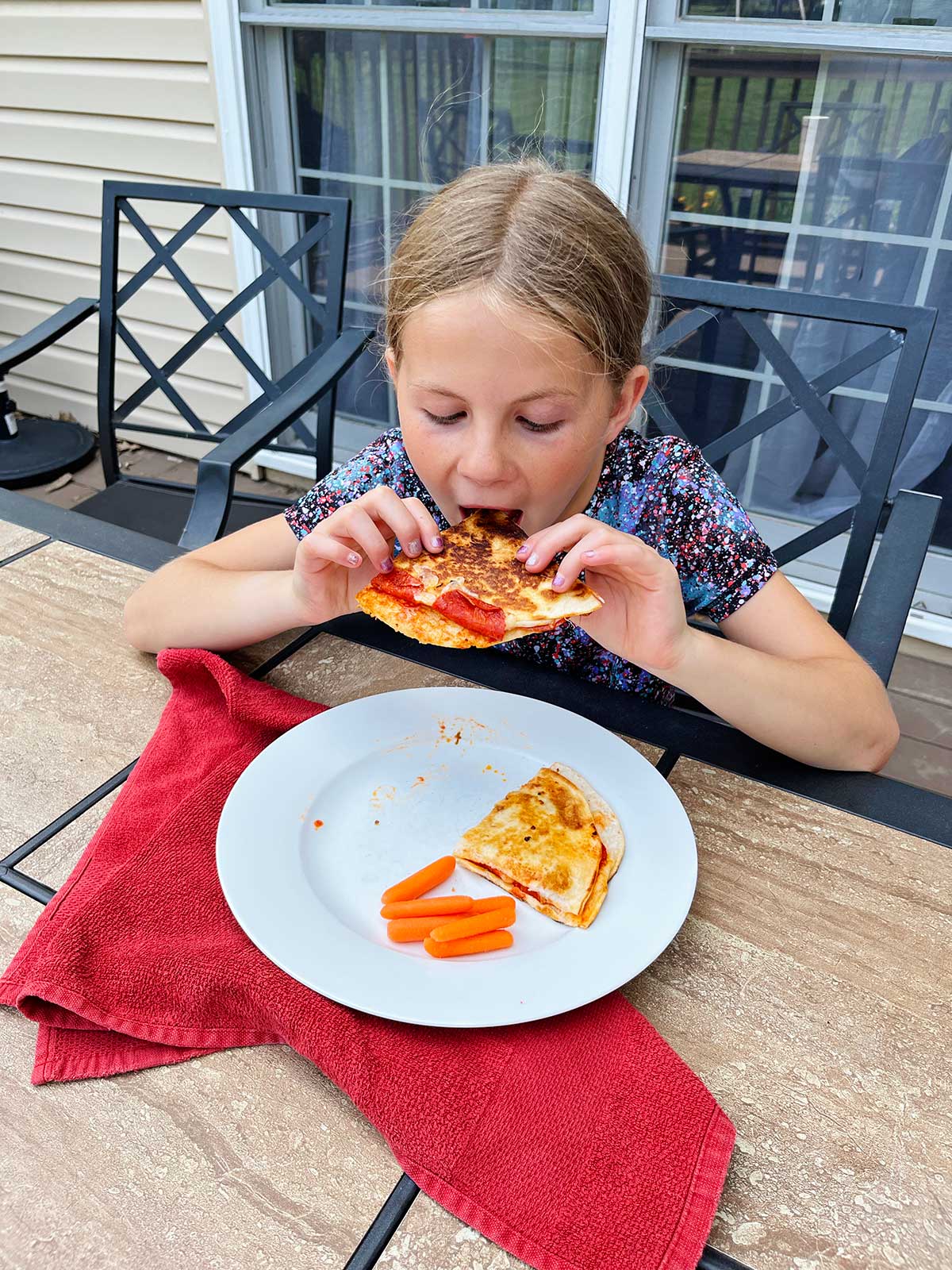 A young girl with blonde hair eating a pizza quesadilla at an outdoor table.