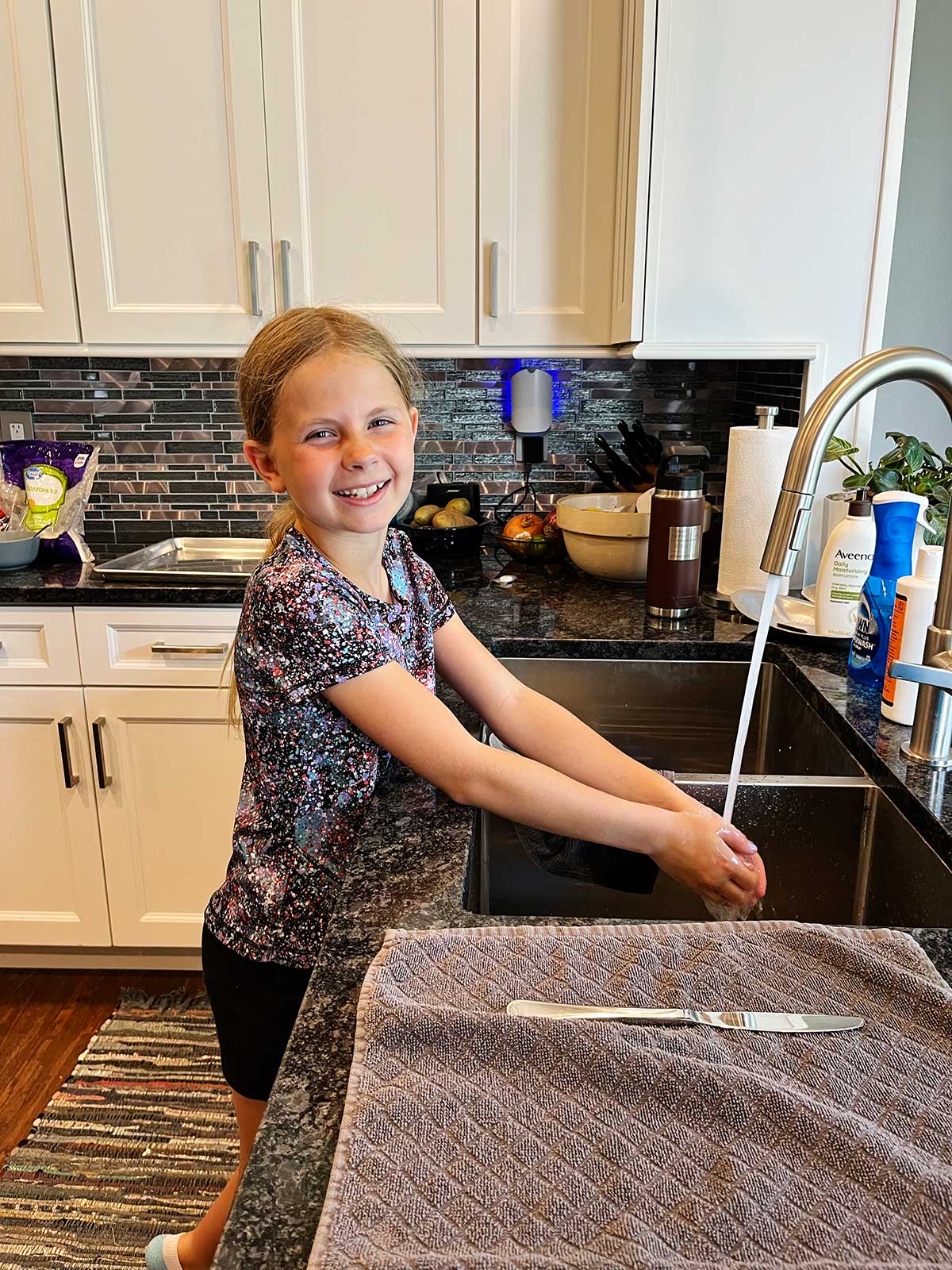 A young girl with blonde hair washing her hands at the kitchen sink.