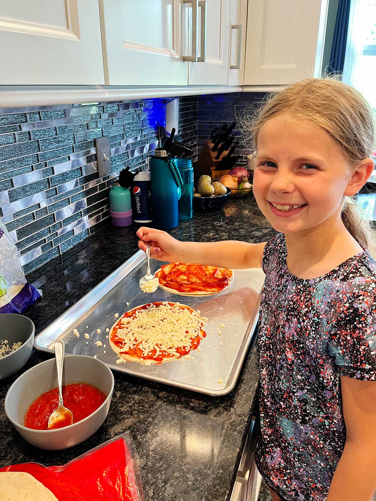 A young girl, smiling at the camera, constructing two pizza quesadillas at the countertop.