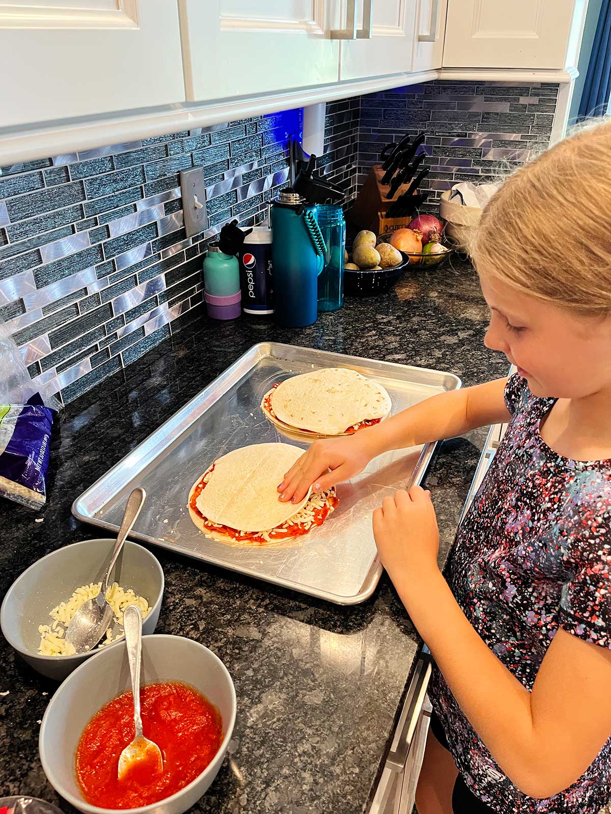 A young girl constructing two pizza quesadillas at the countertop.