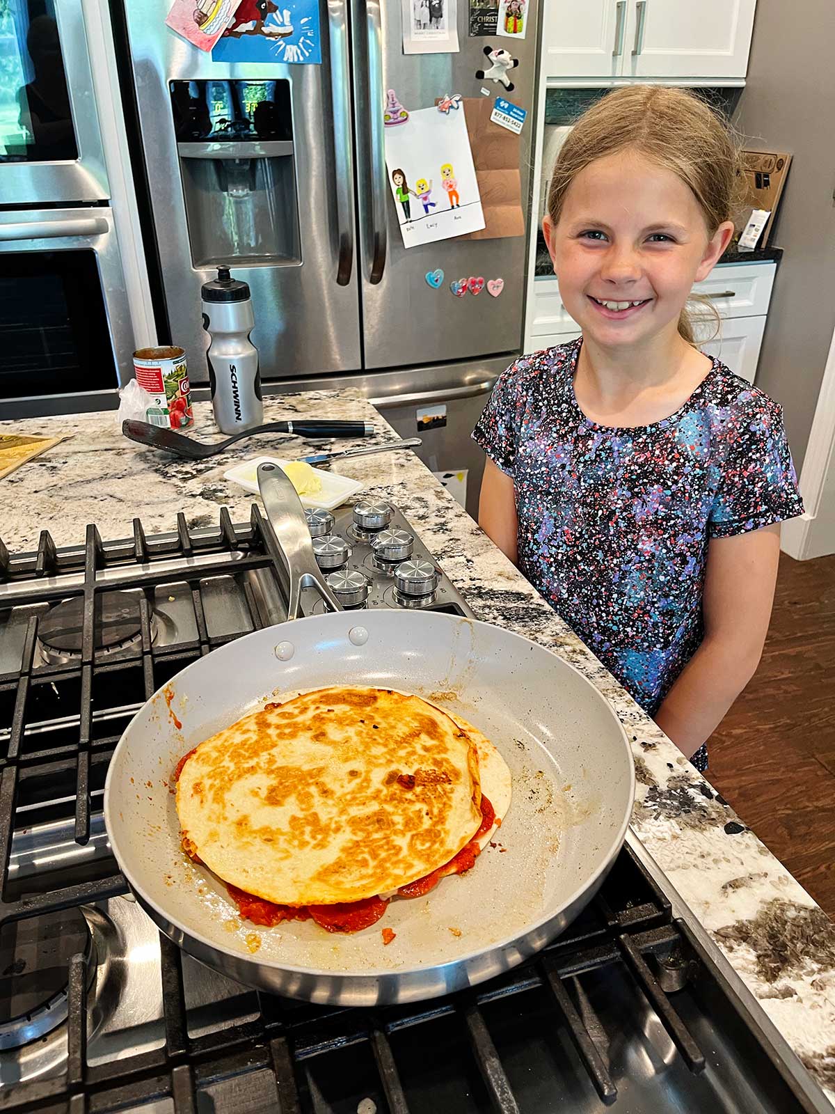 A young girl with blonde hair smiles at the camera while cooking a pizza quesadilla at the stove.