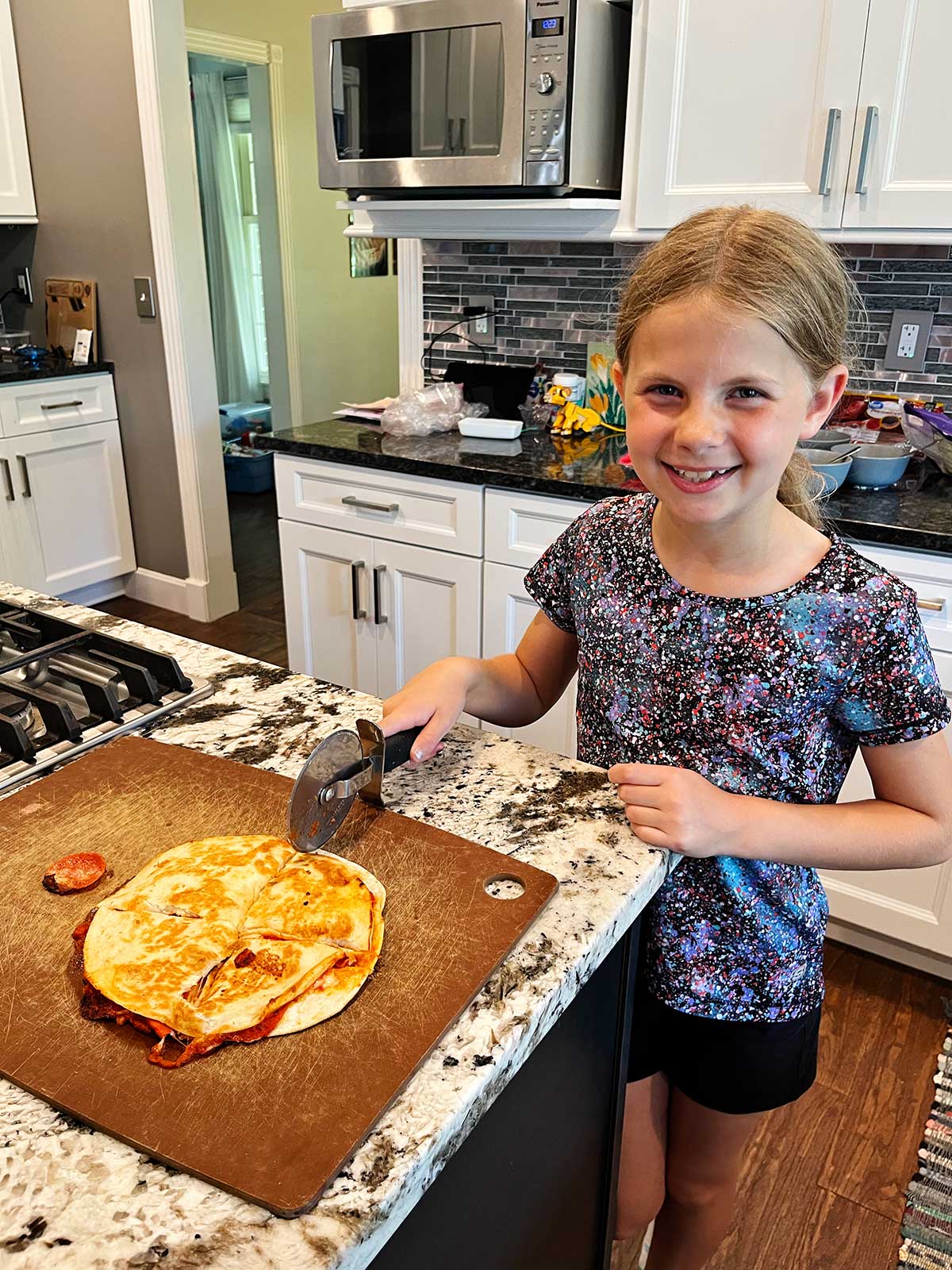 A young girl with blonde hair smiling at the camera and cutting a pizza quesadilla into fourths in the kitchen.