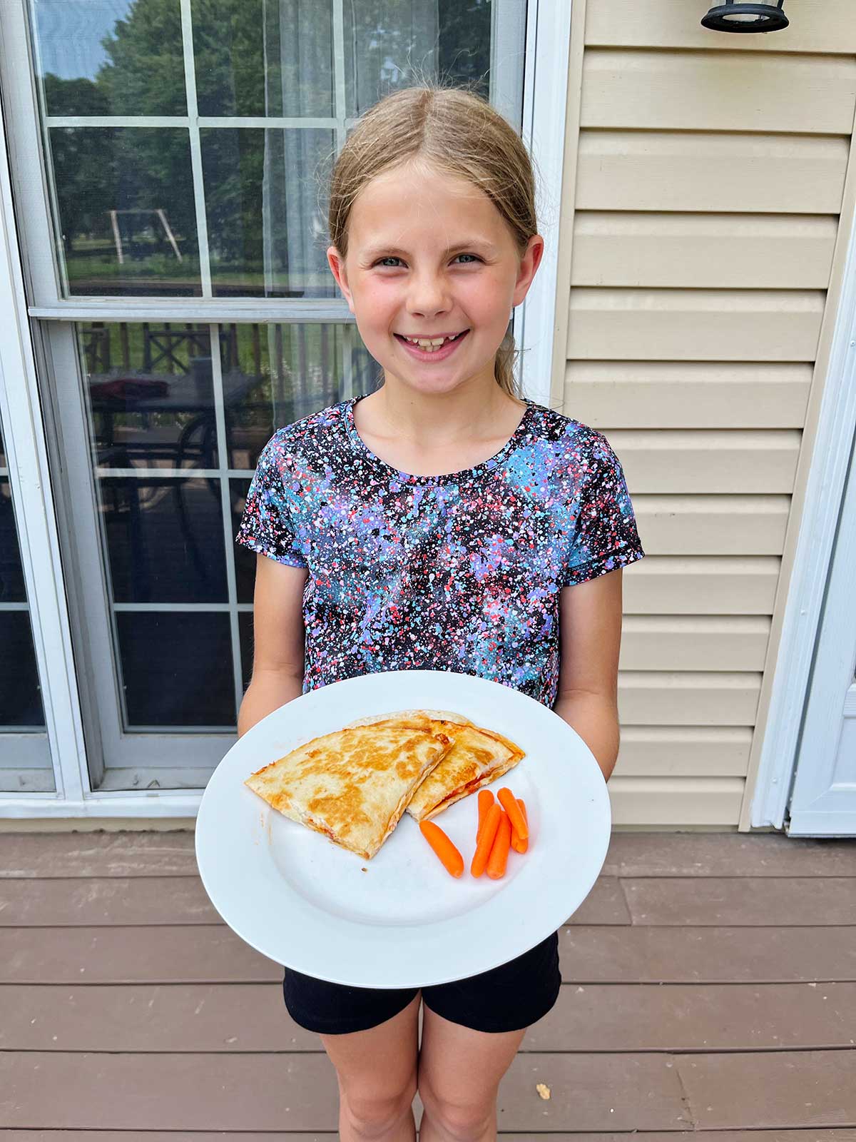 A smiling young girl with blonde hair holding a white plate with two pizza quesadillas and baby carrots outside.