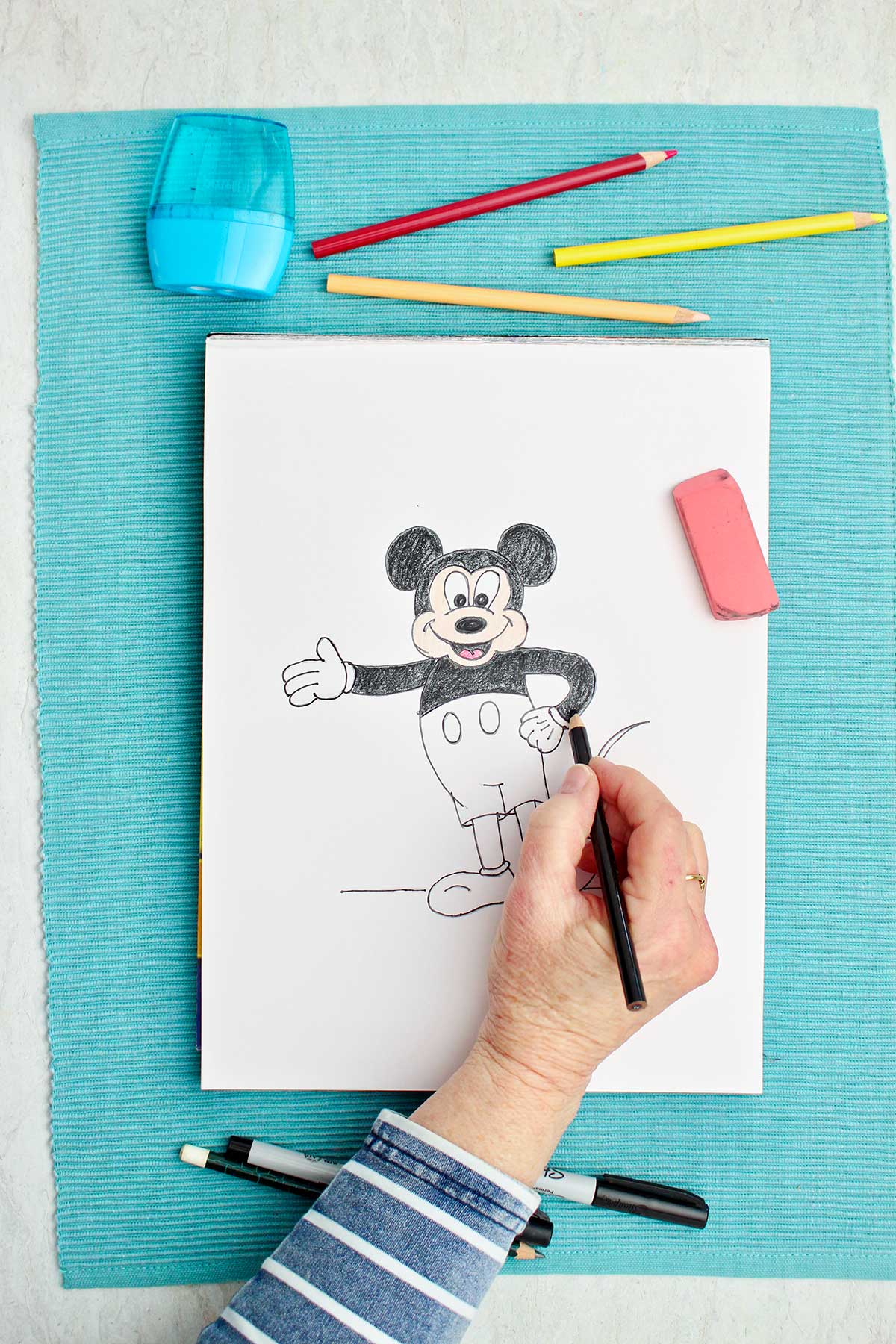 A person coloring in Mickey with a black colored pencil.