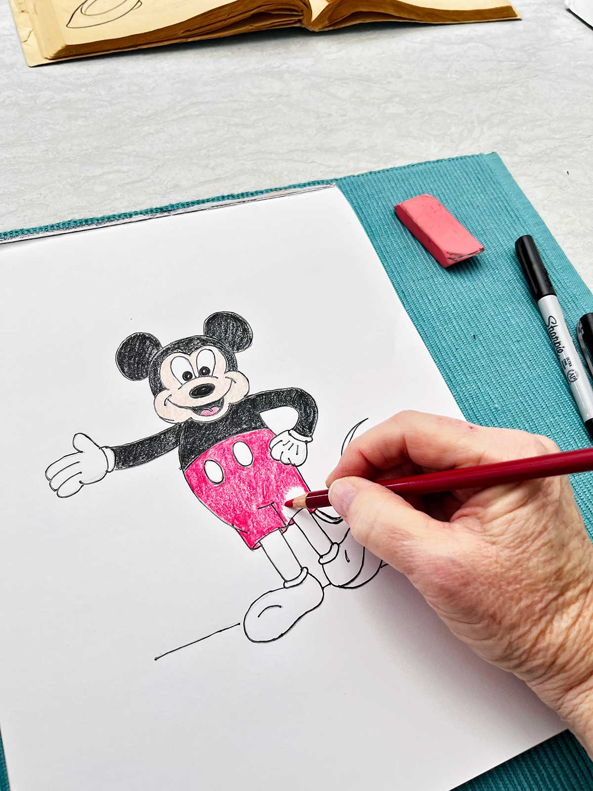A person coloring in Mickey's shorts with red colored pencil.