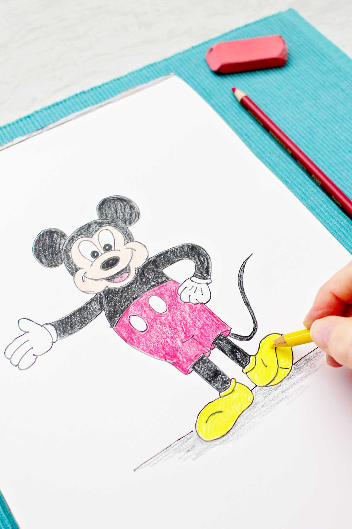 A person coloring in Mickey's shoes with a yellow colored pencil.
