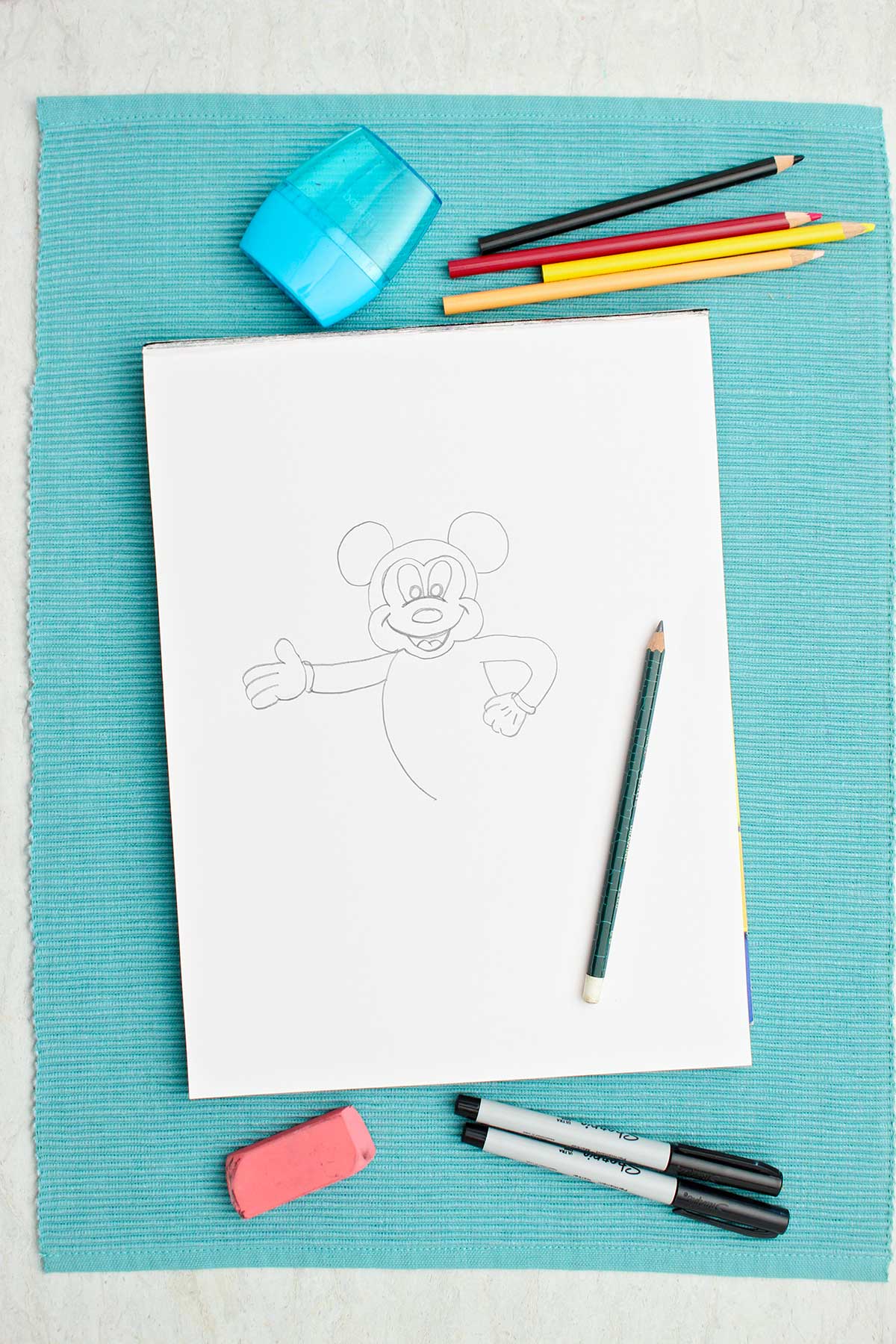 A half completed pencil sketch of Mickey Mouse and some colored pencils and supplies near by.