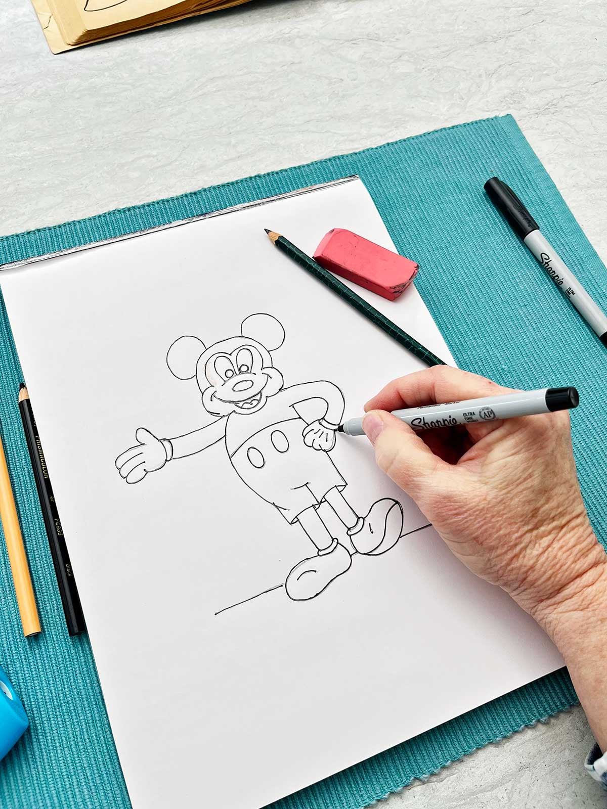 A person going over the pencil sketch of Mickey Mouse with a sharpie marker.