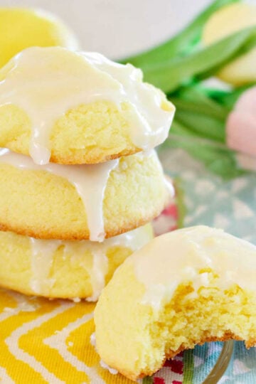 Three lemon cake cookies stacked on a colorful placemat and one cookie with a bite missing.