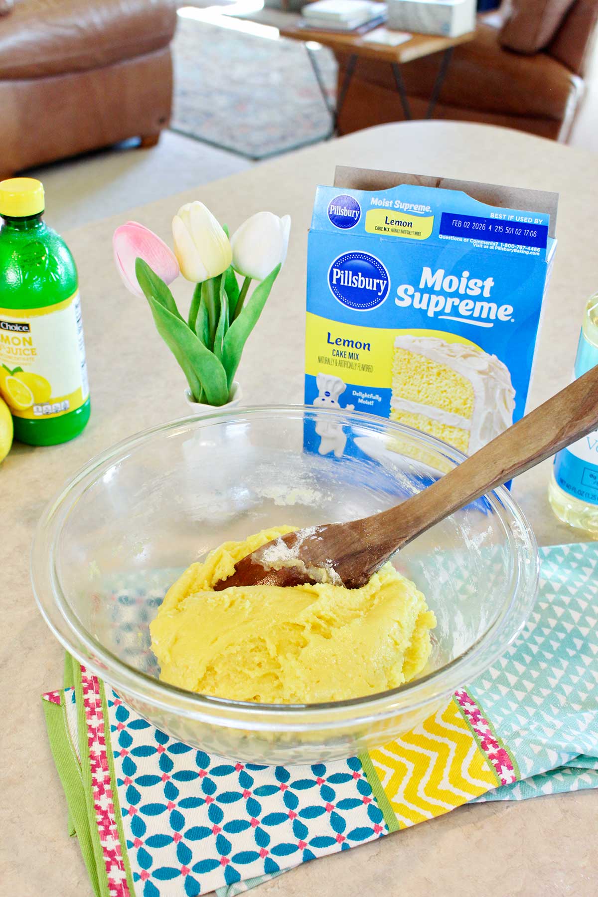 Stirred lemon cake dough in a bowl with supplies and tulips in the background.