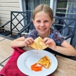 A young girl with blonde hair smiling at the camera and holding a pizza quesadilla at an outdoor table.