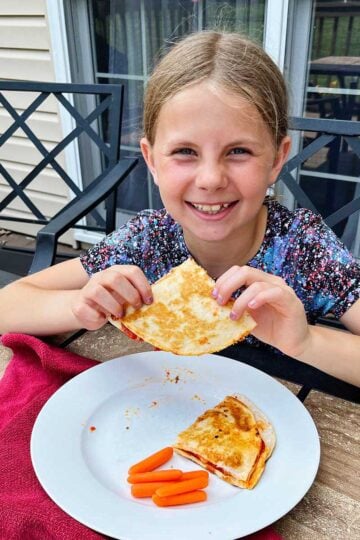 A young girl with blonde hair smiling at the camera and holding a pizza quesadilla at an outdoor table.