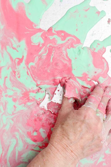 A person picking up a semi solid piece of pink and green oobleck from a cookie sheet.