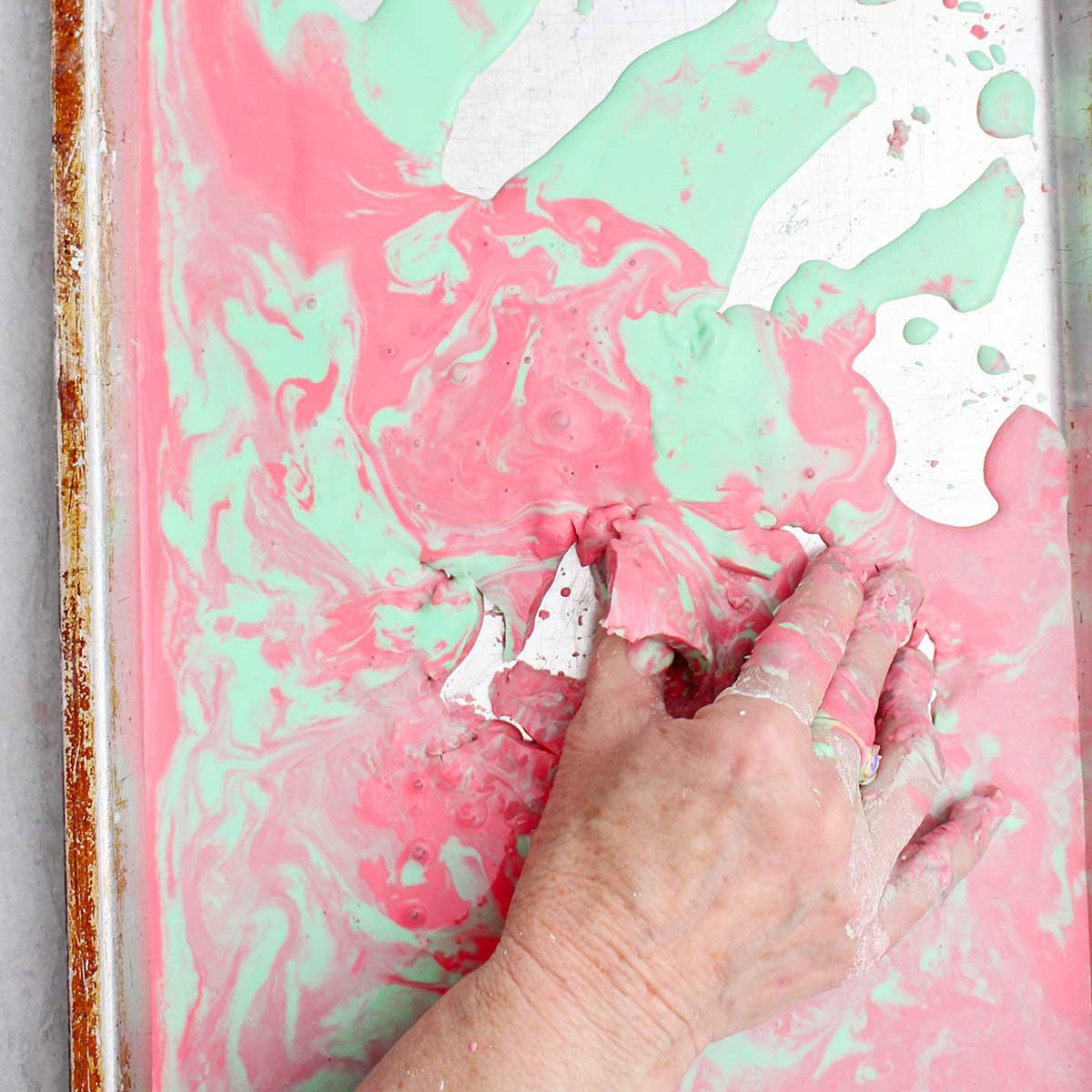 A person picking up a semi solid piece of pink and green oobleck from a cookie sheet.