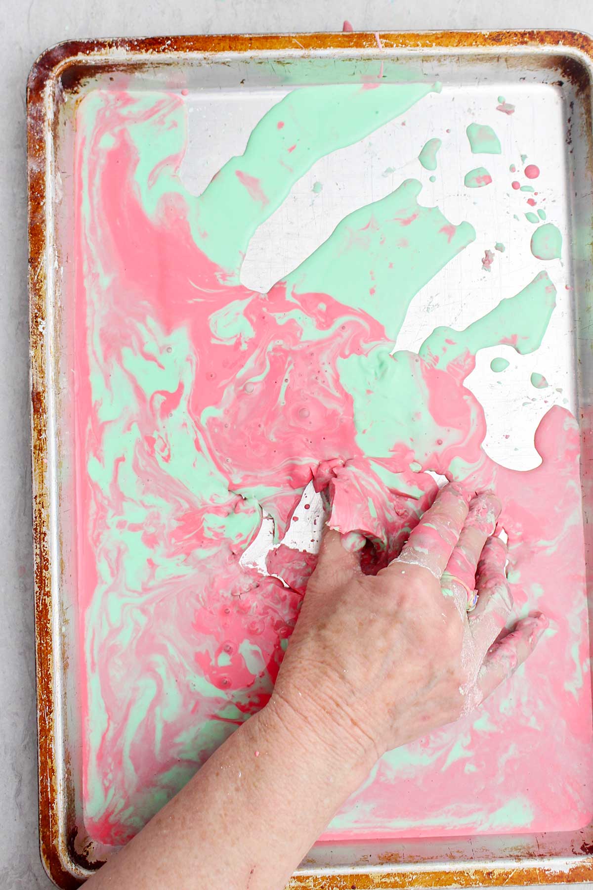 A person picking up a semi solid piece of pink and green oobleck from a cookie sheet.