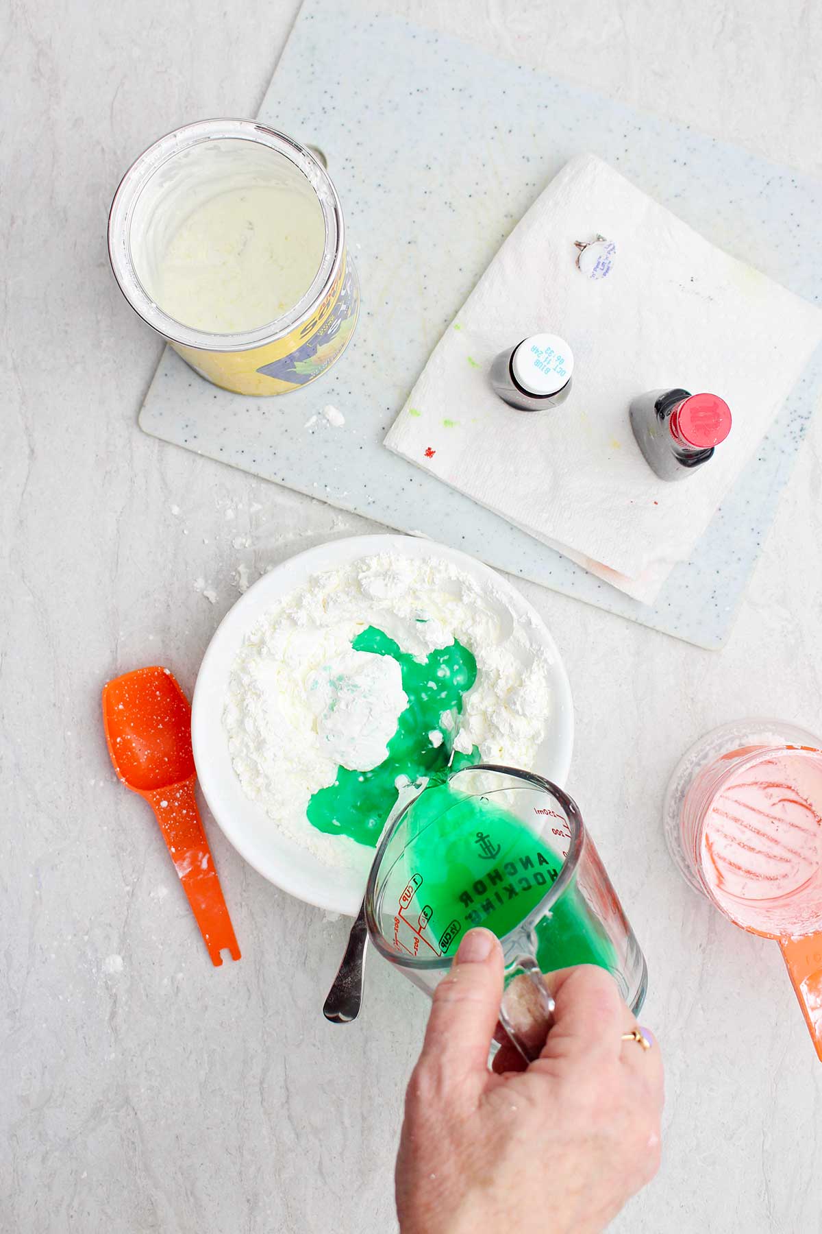 A person pouring water and green food coloring from a measuring cup into a bowl of cornstarch.