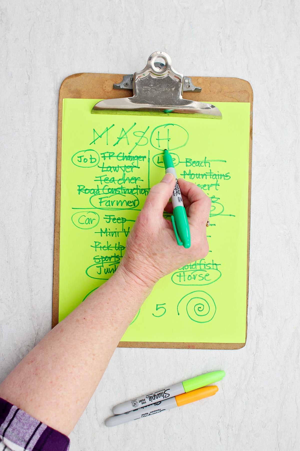 A person circling the "H" in mash with green sharpie on a MASH game.