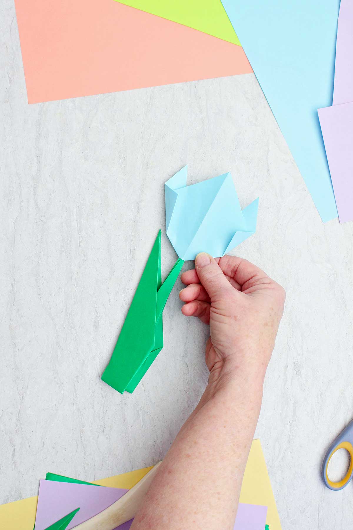 A person holding a completed light blue origami flower with other colorful papers near by.