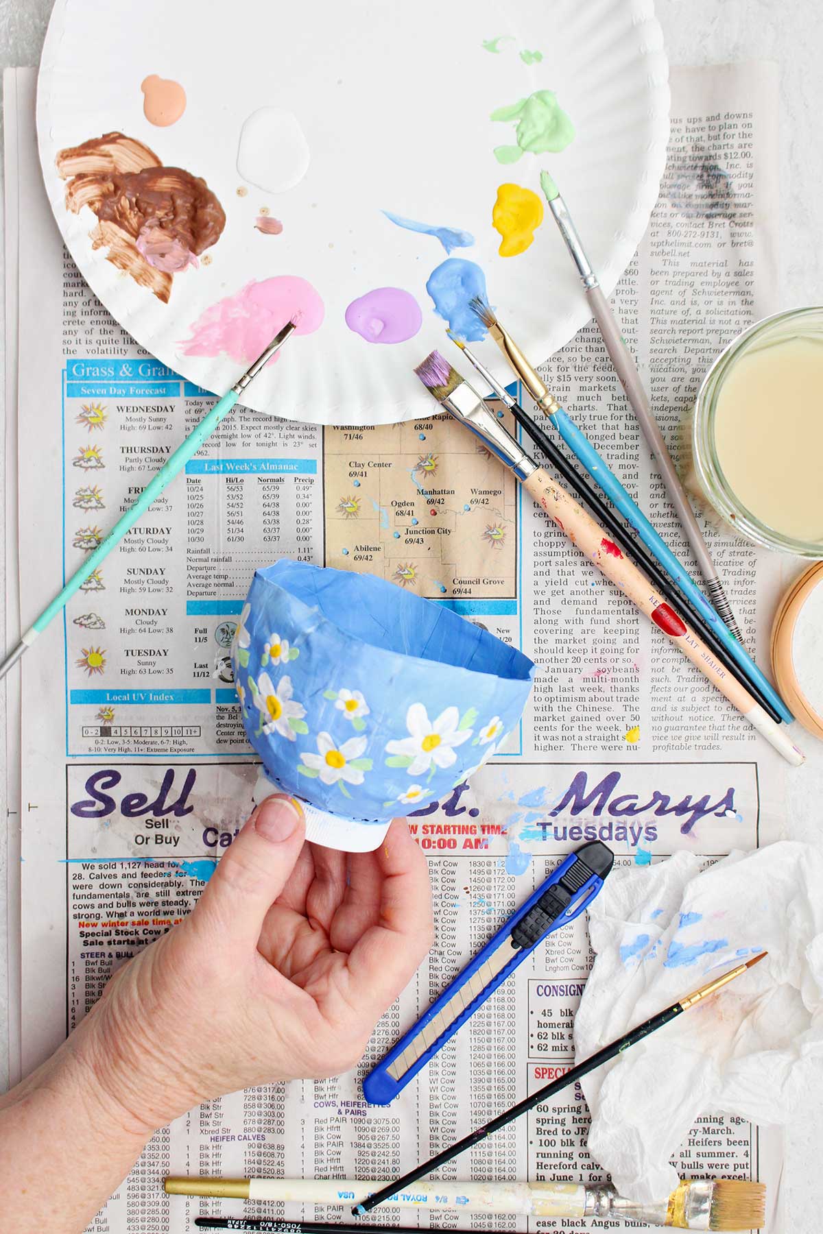 A person holding a paper mache project painted with a light blue background and white and yellow flowers.