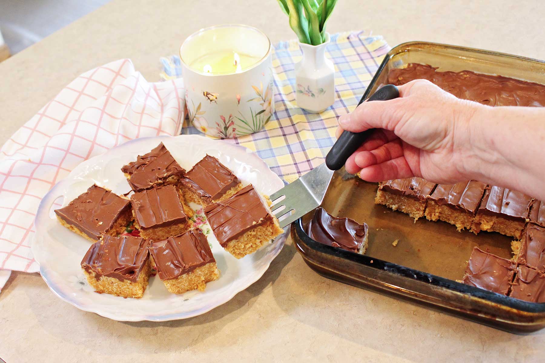 A person serving a scotcharoo on to a plate with some checkered dish towels and flowers near by.