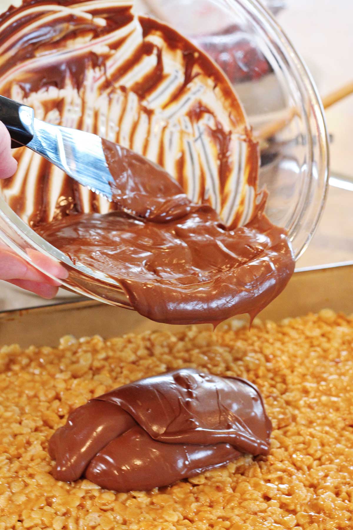 A person pouring melted chocolate out of a bowl onto the scotcharoos.