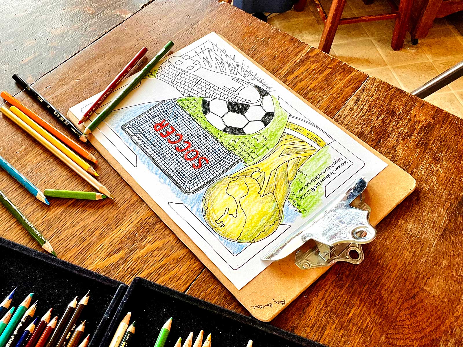 A partially colored in FIFA World Cup coloring page coloring sheet featuring a soccer ball being kicked, a soccer goal, and a FIFA World Cup trophy on a wooden table with colored pencils scattered around.