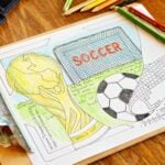 A partially colored in World Cup coloring page coloring sheet featuring a soccer ball being kicked, a soccer goal, and a FIFA World Cup trophy on a wooden table.