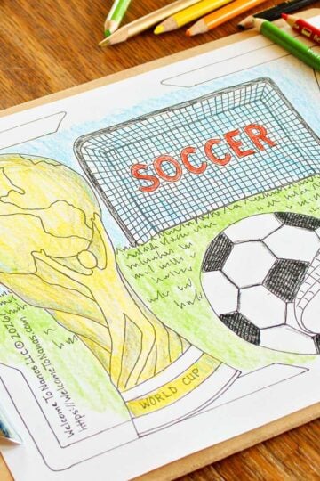 A partially colored in World Cup coloring page coloring sheet featuring a soccer ball being kicked, a soccer goal, and a FIFA World Cup trophy on a wooden table.