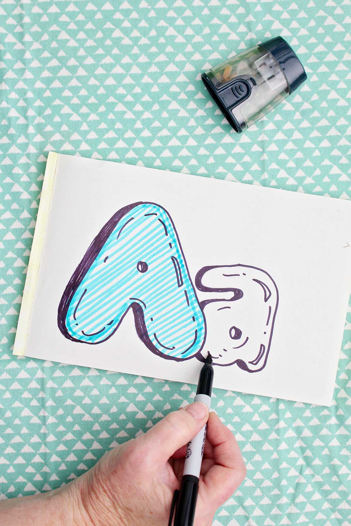 A person adding detail to the bubble letter "a" with sharpie marker.