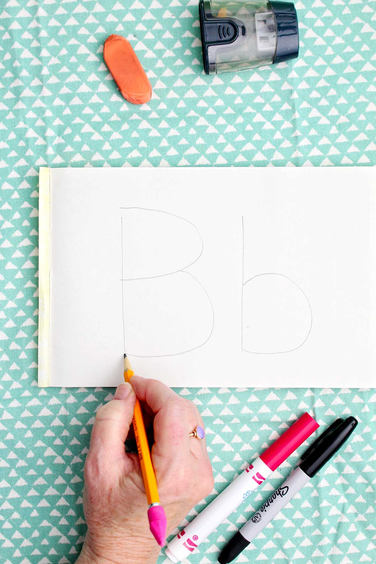 A person writing an upper and lowercase "b" with pencil.