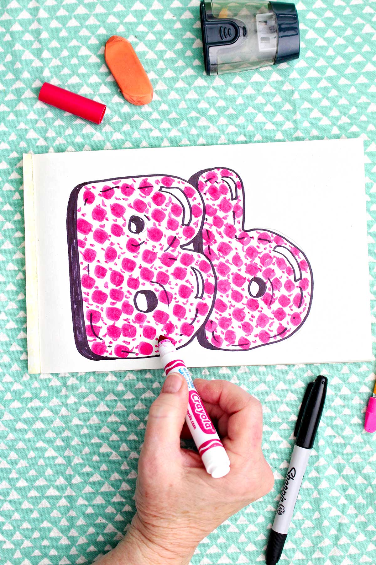 A person coloring in finishing details on the pink pattern inside the "b" bubble letters.
