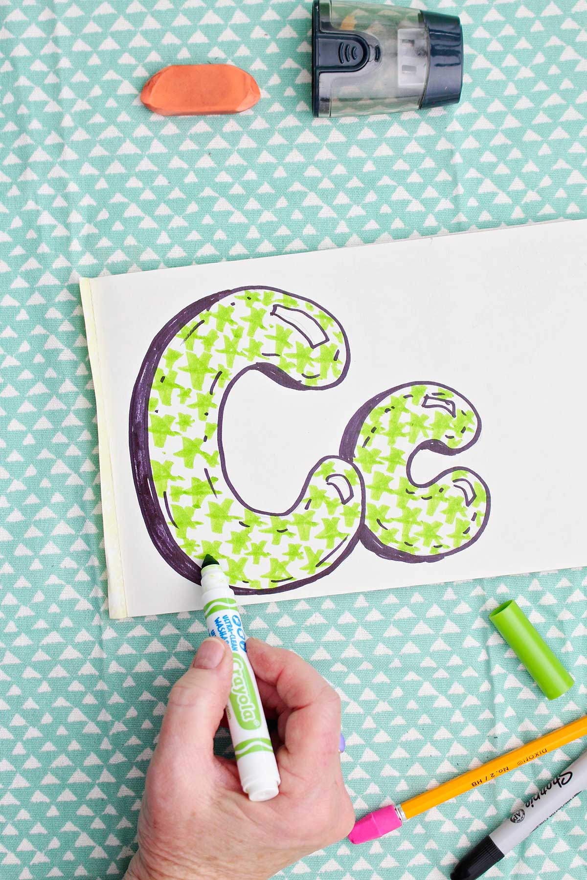 A person coloring in finishing details on the green star pattern inside the "c" bubble letters.