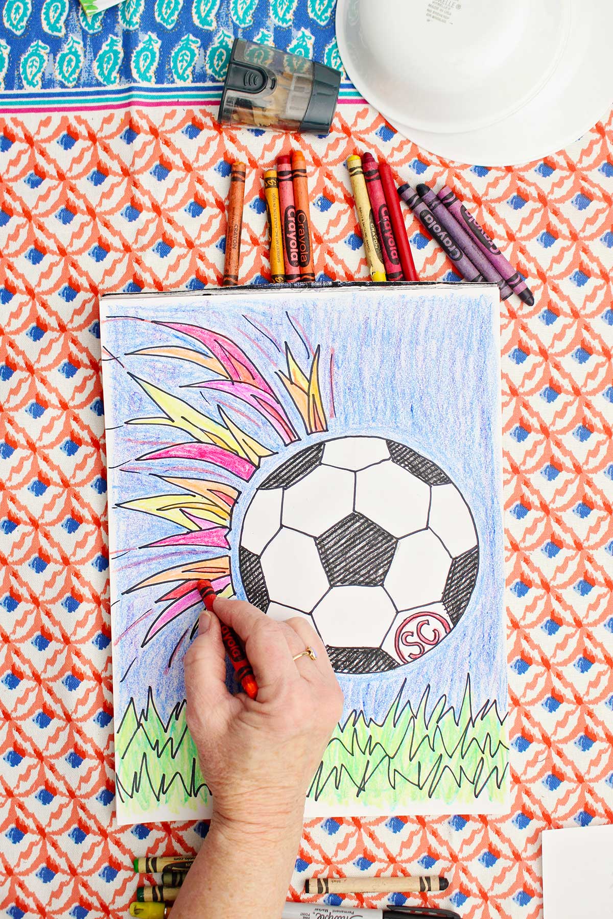 A person coloring in flames on a colored in drawing of a soccer ball flying towards the grass with colorful flames behind it.