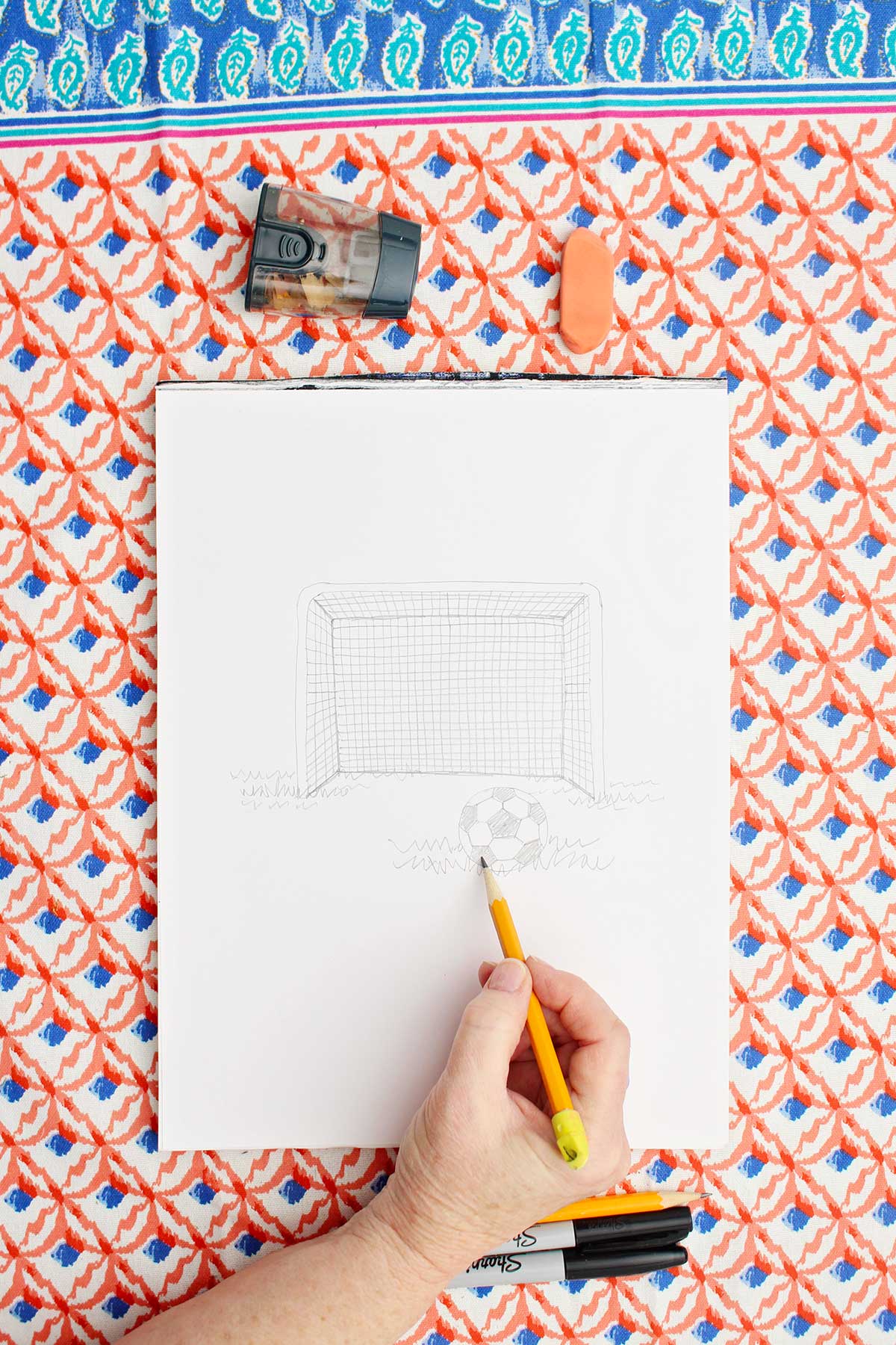 Person sketching a picture of a soccer goal and ball with pencil.