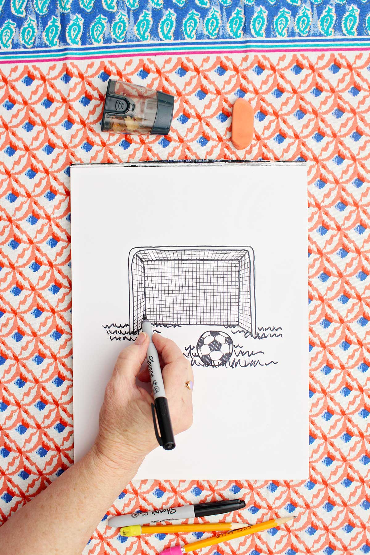 Person going over the pencil drawing of a soccer goal and ball with sharpie marker.