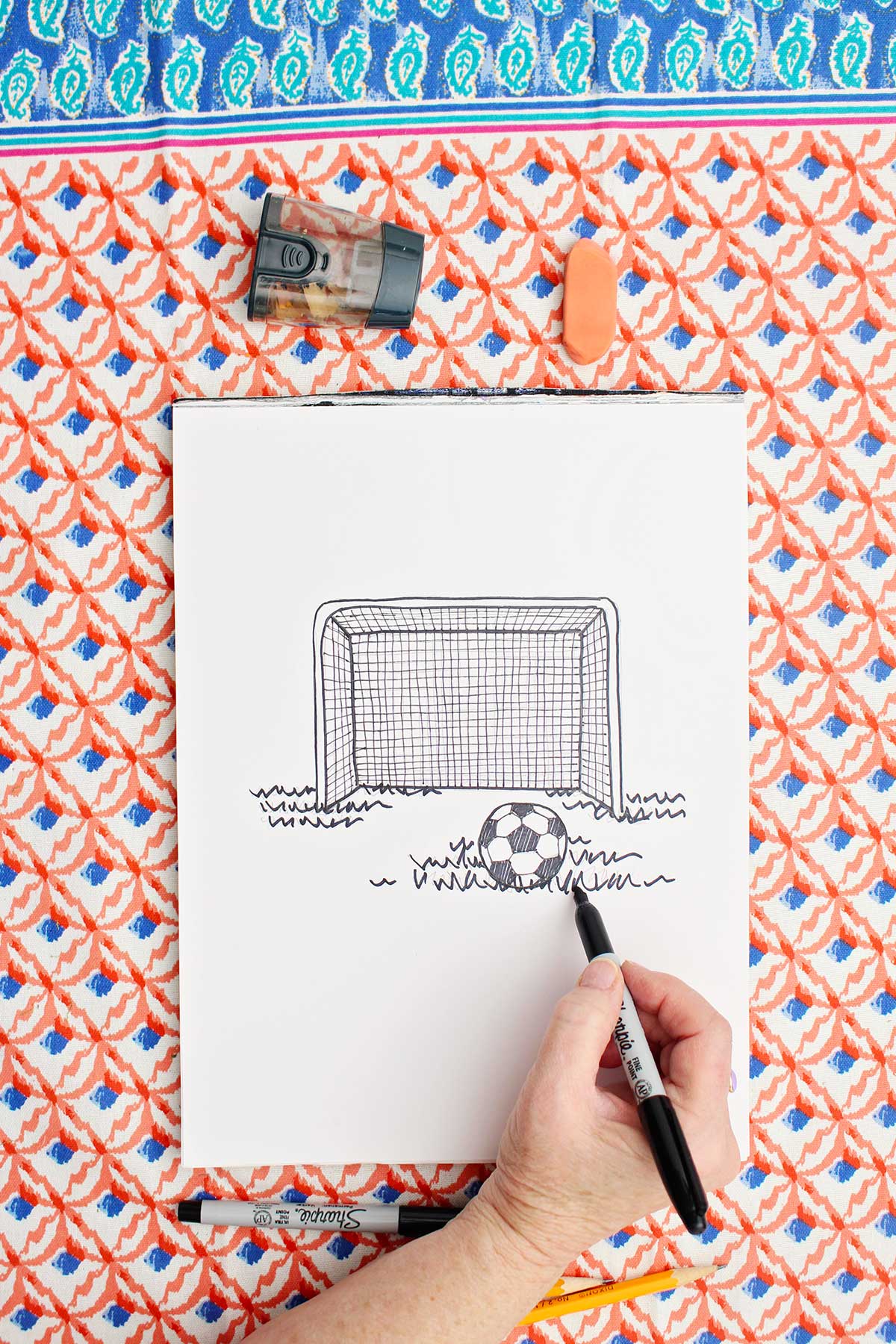Person going over the pencil drawing of a soccer goal and ball with sharpie marker.